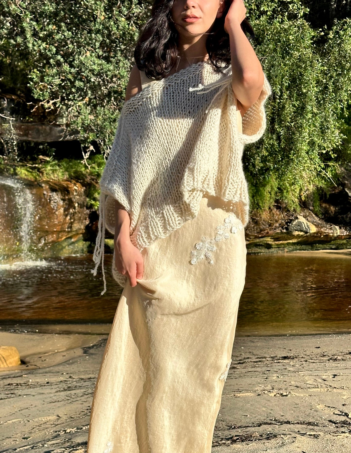 Eleanor silk dress, creme silk dress with lace applique, dress on nora with knitted pullover on top front-view with waterfall greenery background